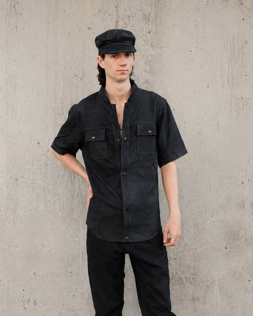 Person wearing a handmade black leather shirt and pants with a cap against a concrete wall.